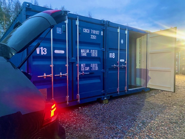 A blue shipping container storage unit partially open, with a vehicle parked nearby and twilight lighting, showcasing its accessibility and storage capabilities.