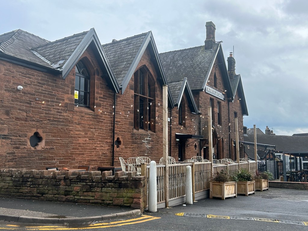 Exterior view of a brick building with large windows and a sloped roof, featuring decorative elements and outdoor seating areas.