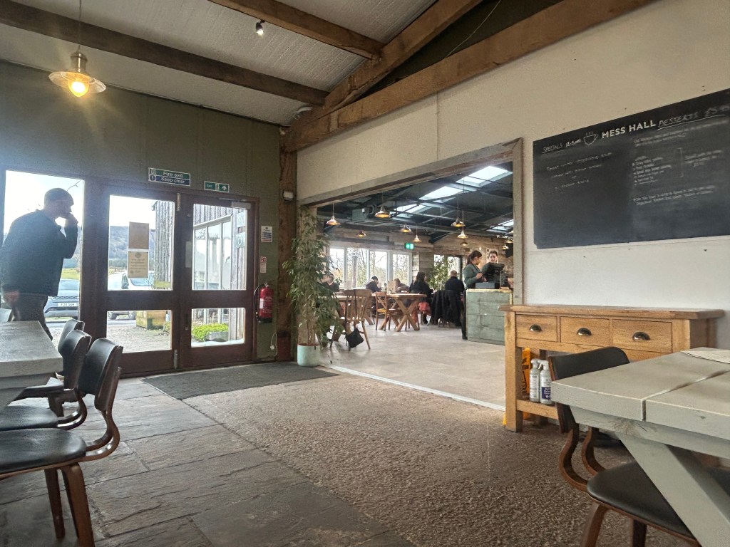Interior view of a cozy café with wooden beams, featuring tables and chairs, a menu board, and patrons dining in a relaxed atmosphere.