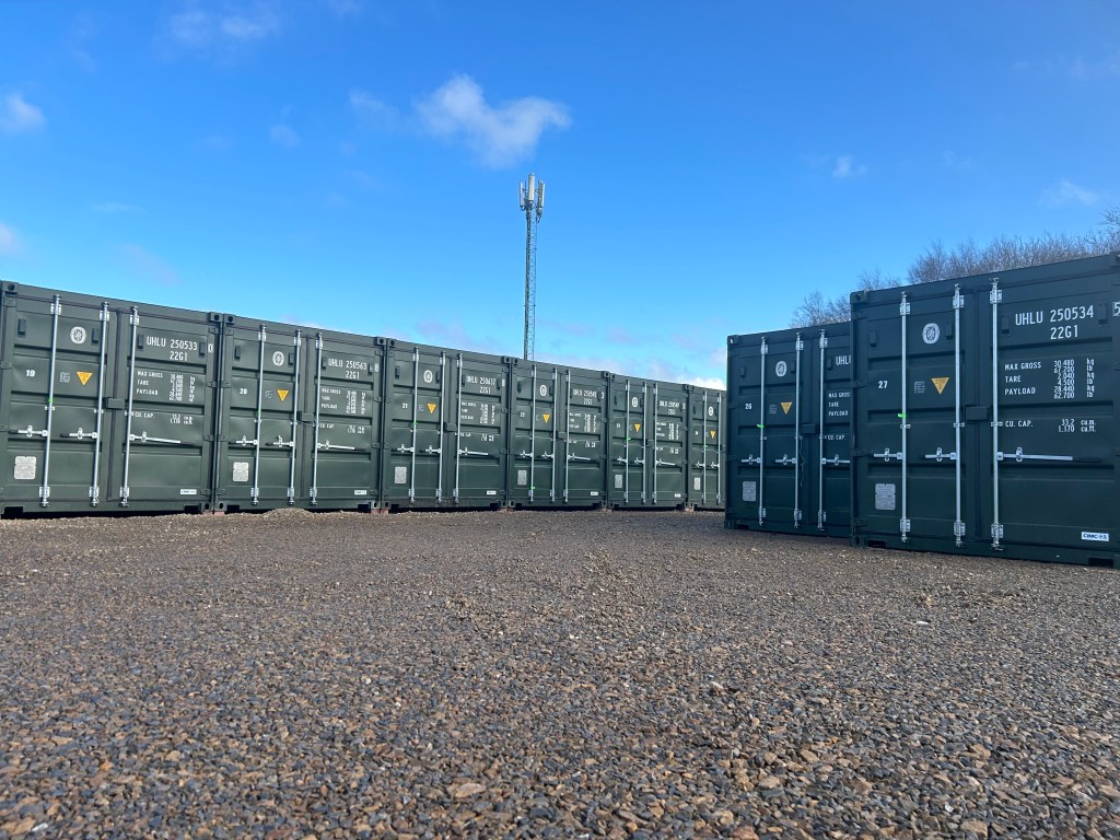 A row of green shipping containers on a gravel surface with a clear blue sky in the background.