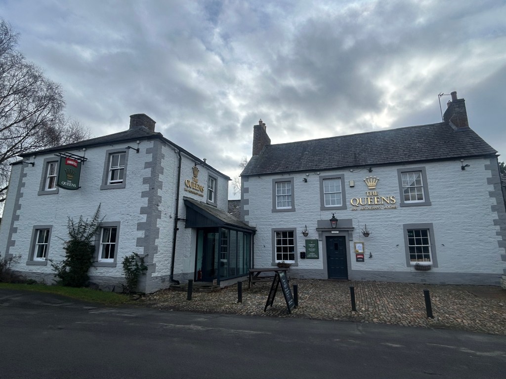 Exterior view of The Queens pub, Warwick-On-Eden, Carlisle, Cumbria featuring two buildings with white walls and grey accents, under a cloudy sky.