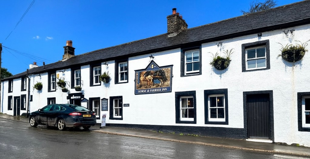 Exterior view of a traditional whitewashed pub named 'Horse & Farrier Inn' with hanging flower baskets, blue sky, and a parked car in front.