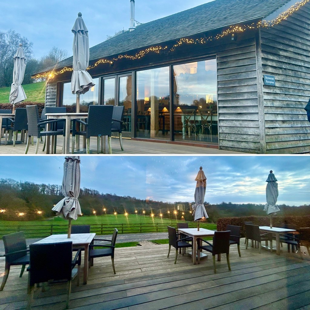 Exterior view of Hidden River Restaurant, Longtown, Cumbria. A wooden restaurant with large glass windows, featuring patio furniture and umbrellas. The setting is scenic with trees and outdoor lighting visible in the background during dusk.