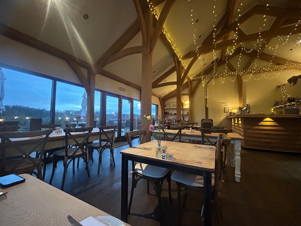 Hidden River Restaurant, Cumbria. Cosy restaurant interior with wooden beams, tables set for dining, and large windows showcasing a dusky sky. String lights hang from the ceiling, creating a warm atmosphere.